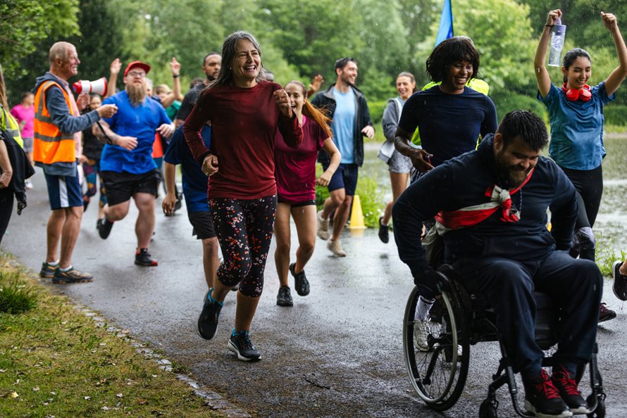 A group of people taking part in a fun run as a community group and it is raining. At the front of the group there is a wheelchair user.
